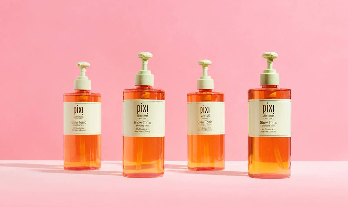 Four Pixi Glow Tonic pump bottles filled with orange liquid, arranged in a row against a solid pink background. View 1