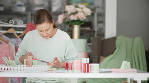 Petra in a light sage green sweatshirt sits at a white table, applying makeup from a small pink tube to her hand. Several cosmetic products in pink, red, and white packaging are arranged on the table in a softly lit room with a blurred background of clothing racks and flowers.