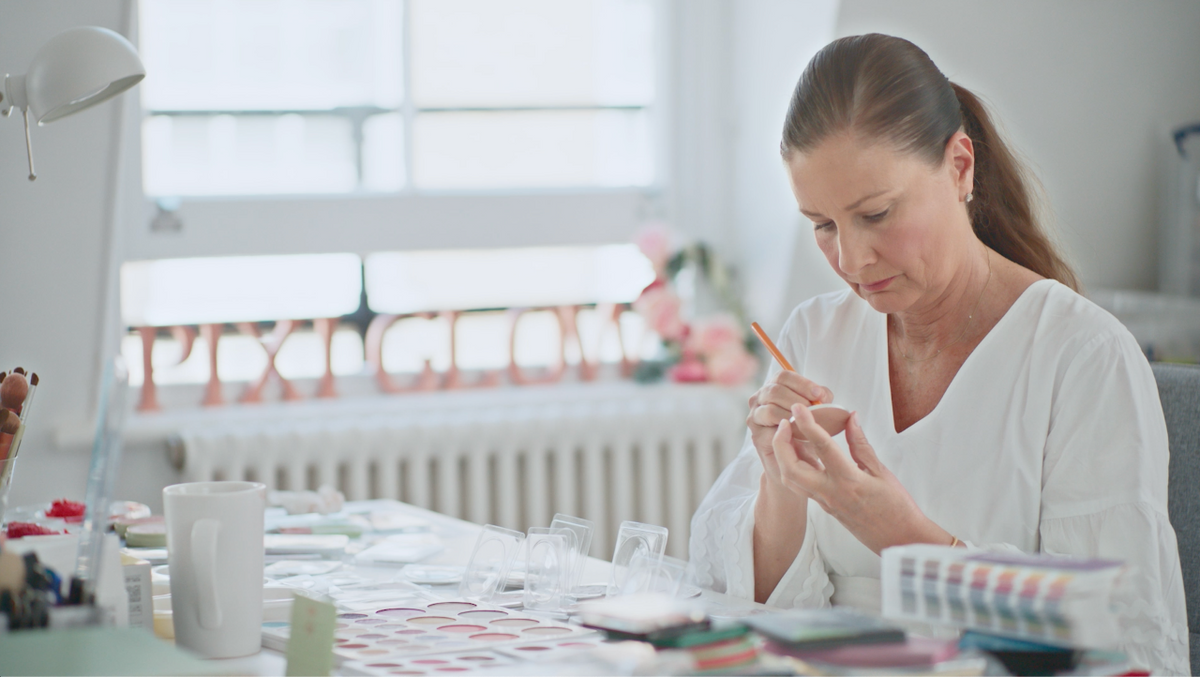 A woman with her hair in a ponytail sits at a table, carefully examining a small clear object with a pencil. The room is bright and airy with a large window in the background, and the table is cluttered with various craft or sample materials.