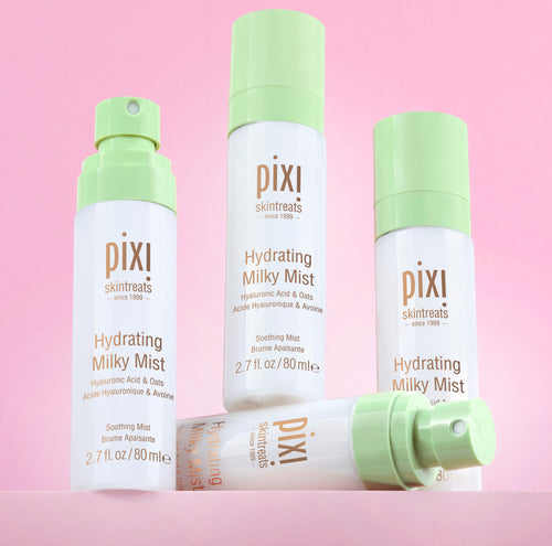 Three Pixi Skintreats Hydrating Milky Mist bottles in white and light green packaging, arranged on a solid pink background with one bottle lying down in front. view 1
